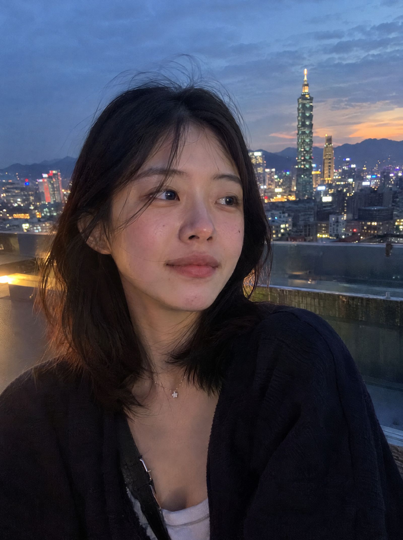 Korean woman with windblown hair, Taipei 101 and a sunset sky behind her, smartphone photo